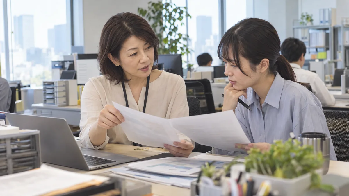 午後のオフィスで、資料を手に同僚と落ち着いて打ち合わせをする40代の女性